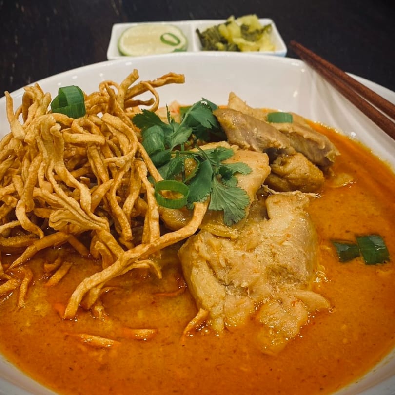 A bowl of Thai Khao Soi with chicken, crispy noodles, cilantro, and green onions, with lime and pickled greens on the side.