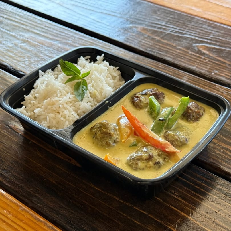 Thai green curry with meatballs, bell peppers, and rice in a black takeout container.